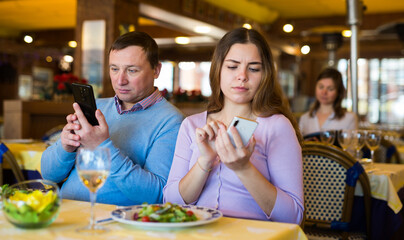 European man and woman sitting at table in restaurant and using their smartphones.