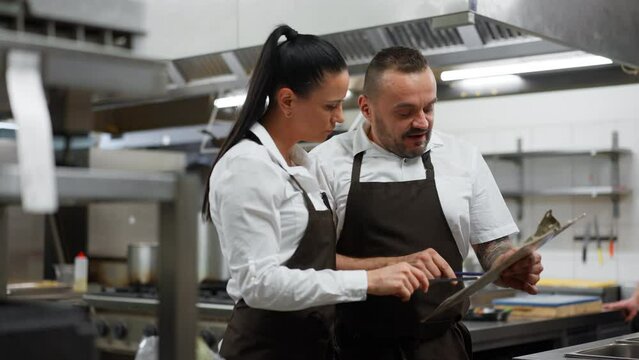 Chef and cook discussing menu indoors in restaurant kitchen.