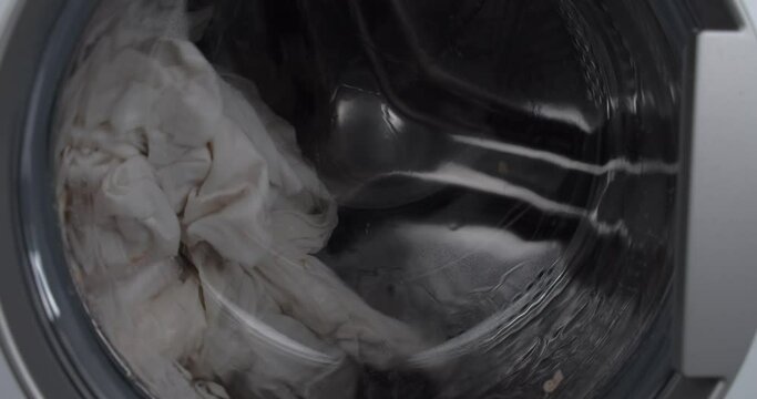 White cloth and water spins inside washing machine drum, close up view