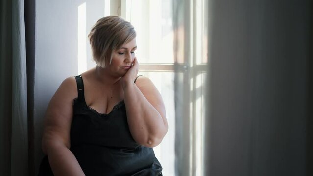 Depressed lonely fat woman sitting aby window at home and thinking.