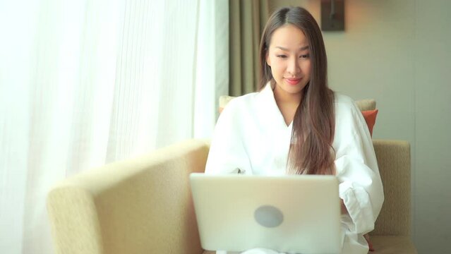 A close-up of a young woman in a bathrobe sitting in a window-side chair as she reads her laptop screen.