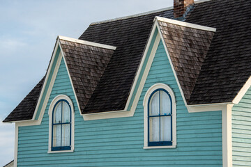 Teal green vintage building with cream trim and dark roof shingles. The top of the building has two dormers and multiple old single hung windows. The background has blue skies with white clouds. 