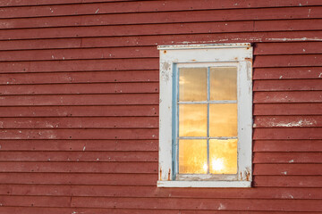The exterior of an old red country wooden shed exterior wall with a white framed closed glass single hung window. In the window is a reflection of the cloudy sky with the orange-colored sun setting. 
