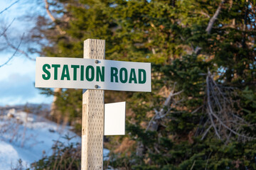 A white metal street name sign is attached to a square wooden post. The green text on the sign spells station road in capital letters. There are large green trees and a blue sky in the background. 