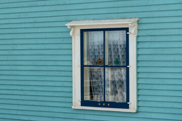 The exterior outside wall of a teal blue heritage wood building with dark green and tan trim. The country style house has narrow clapboard and a single double hung window with lace curtains hanging. 