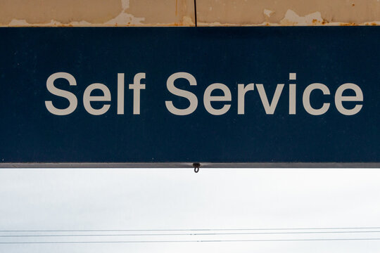 A Black And White Sign With The Text Self Service. The Sign Backing Is Black And The Letters Are White In Color. The Sign Is Affixed To An Exterior Wall At A Self-service Gas Station Checkout.  