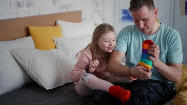 Father Putting On Different Socks To His Little Daughter With Down Syndrome When Sitting On Bed At Home.