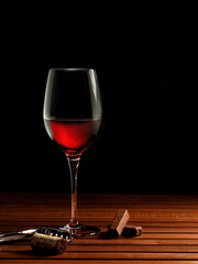 A glass of red wine on a wooden table top with two pieces of chocolate bar and corkscrew