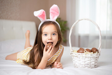 Happy Easter. A cute girl with rabbit ears eating chocolate eggs on a bed in a bright bedroom....