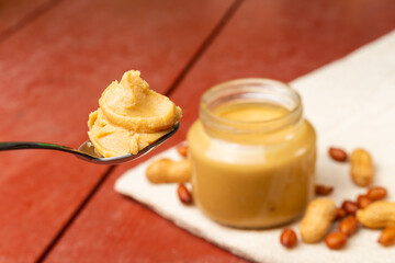 Spoonful with peanut butter, and jar in the background with peanuts on the wooden table.