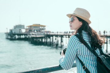 side view portrait of asian chinese woman on seafront leaning on railing enjoy sun flare. Young charming girl in hat and sunglasses on pier holding camera enjoy summer holidays. female backpacker.