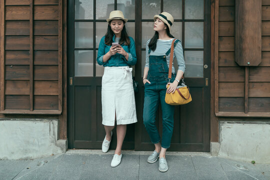 full length elegant girl tourists standing by wooden door in front of old japanese traditional house waiting. young woman using mobile phone while friend carrying bag looking around beside outdoors