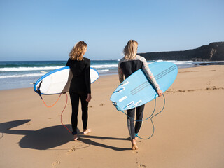 2 caucasian surfer women on the beach walking to the waves. They are wearing winter wetsuits.