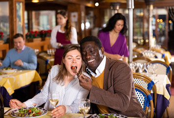 Positive adult biracial family couple having fun during dinner in cozy restaurant. Cheerful african american man feeding his european wife with fresh vegetable salad