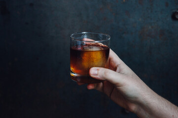 hand holding rocks glass of whiskey with ice against dark blue metal background. copy space. 