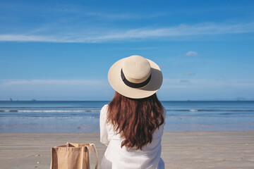 Fototapeta na wymiar Rear view image of a woman with hat and bag sitting on the beach with blue sky background