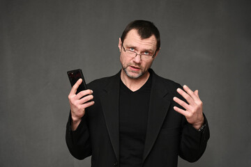 A businessman in a jacket and glasses with a smartphone in his hand is talking to the camera. Portrait of an adult man on a gray background in the studio