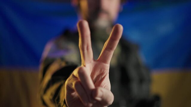 Soldier in military uniform showing peace sign with Ukrainian flag in background.