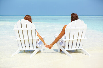 Loving couple relaxing on deck chairs. Rear view of loving couple holding hands while relaxing on deck chairs at seashore. - Powered by Adobe