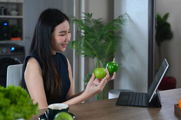 Happy young woman reading recipe from digital tablet while preparing vegetable salad in kitchen.