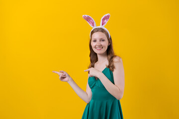 Young caucasian girl wearing bunny ears on yellow background wearing green dress, pointing to the side with open palms, showing copy space, presenting advertisement smiling, excitedly happy