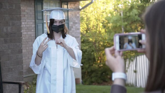 High school girl wearing cap and gown with face mask 2020 as she throws her hands up while she has her photo taken.