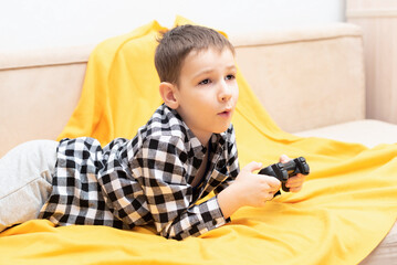 The child boy in checked shirt lying on the couch with black joystick in his hands playing the...