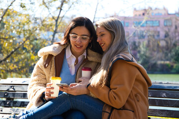 A lesbian couple sitting on a bench checking their social networks over a cup of coffee. The women are young and recently married. Concept lgtb, gays and lesbians. Rights and equality