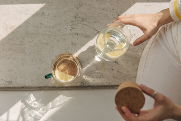 Top view of glass with drink near jug of lemonade in sunlight. Detail of body part of young fair-skinned girl holding wooden cover. Windowsill breakfast concept