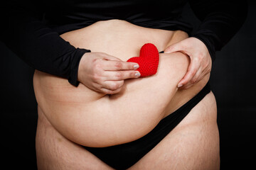 a woman holds a red heart in her hands on a fat belly on a black background. obese person. body positivity concept