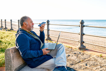 Bearded mature man with laptop working outdoors while sitting on bench at seaside. Modern lifestyle, connection, business, freelance work concept. Smiling gray-head man talking on the smartphone