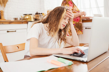 a tired schoolgirl girl is doing homework at home in the kitchen with a laptop
