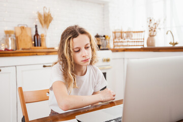 a tired schoolgirl girl is doing homework at home in the kitchen with a laptop