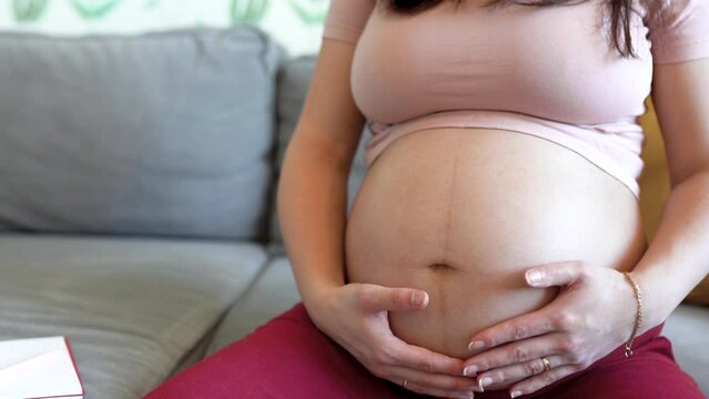 Pregnant woman gently touches her big belly while sitting on the sofa in the living room