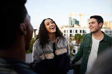 Biracial female laughing with group of multi-cultural friends socializing on a rooftop terrace at dusk.
