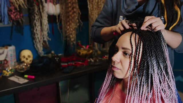 Unrecognizable person makes hairstyle for young woman in salon. Professional hairstylist makes multicolored senegalese pigtails for client.