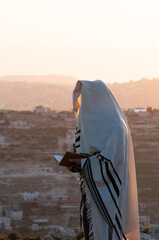 A Jewish man holding a siddur prayerbook and wearing a tallit and tefillin prays the morning...