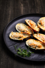 baked mussels with cheese, on a dark plate with herbs and lemon on a wooden background