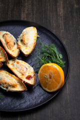 baked mussels with cheese, on a dark plate with herbs and lemon on a wooden background