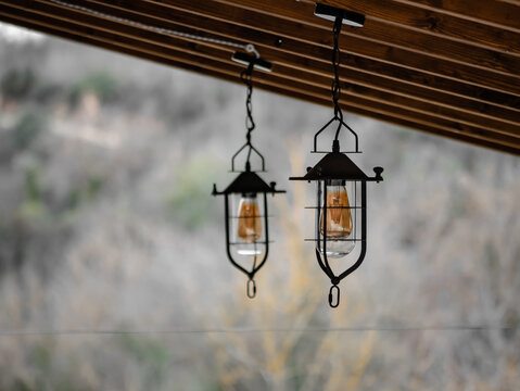 Two lanterns with large orange bulbs hang from the wooden porch of a rural town house and the forest vegetation out of focus in the background