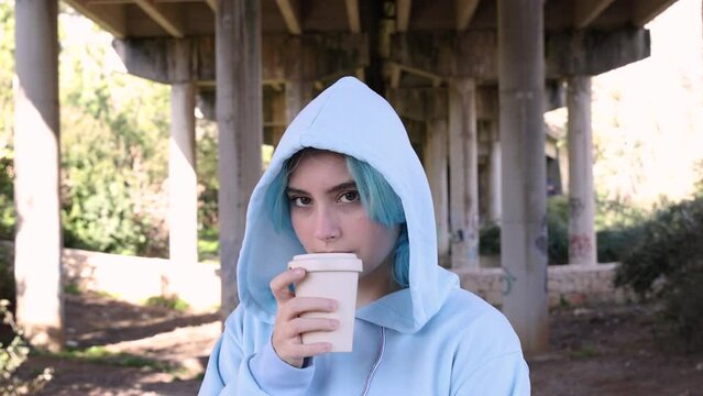 Teenage girl in blue oversize hoodie drinking coffee to go. Blue haired teen girl staying outdoors against bridge pillars. Mockup, head shot. Hipster and adolescence concept
