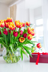 Huge spring bouquet of tulips in vase and gift box on the table with light classic interior design background. Gift for holiday, birthday, 8 March, Mother's Day, Valentine's day, Women's Day. Vertical