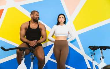 Athletic couple: adult afro american man and young adult caucasian woman take a break and talking near the cycling machine in a sports club.