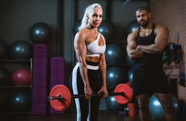 Fit young caucasian woman lifting barbell, working out in a gym with afro-american personal trainer assistance cheering her on. Personal training and bodybuilding concept.