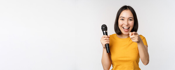 Enthusiastic asian girl with microphone, pointing finger at camera, suggesting you to sing,...