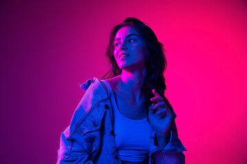 Beautiful young emotional girl, student with long dark hair isolated on gradient pink-blue background in neon light. Concept of emotions, youth