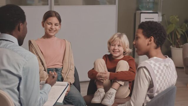 Medium slowmo shot of three diverse cheerful school kids talking to African-American psychologist during group therapy session