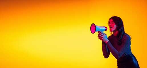 Beautiful young excited girl, student shouting at megaphone isolated on gradient yellow background...