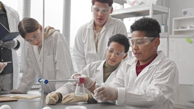 Medium slowmo of Biracial schoolboy in lab coat making chemical experiment by putting brown powder and transparent liquid into glass flask and having lab smoke from flask as reaction