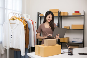 Young beautiful asian woman business owner at fashion store holding laptop check stock on rails clothing. Asian female entrepreneur working in clothes shop.